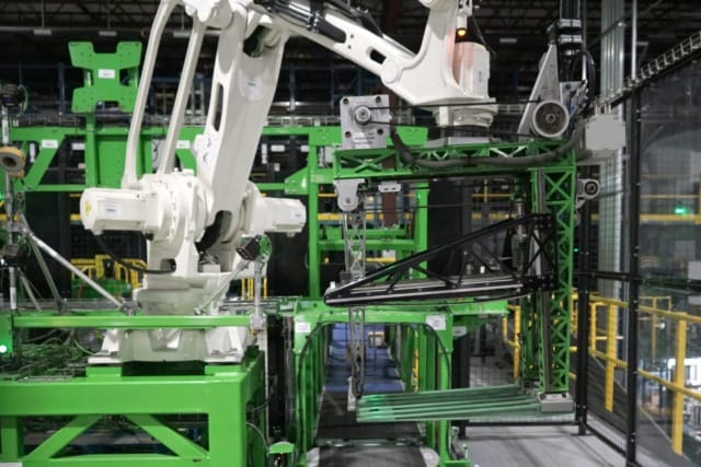 Brazo robótico industrial en funcionamiento en el entorno de una fábrica moderna, rodeado de estructuras metálicas verdes y maquinaria.