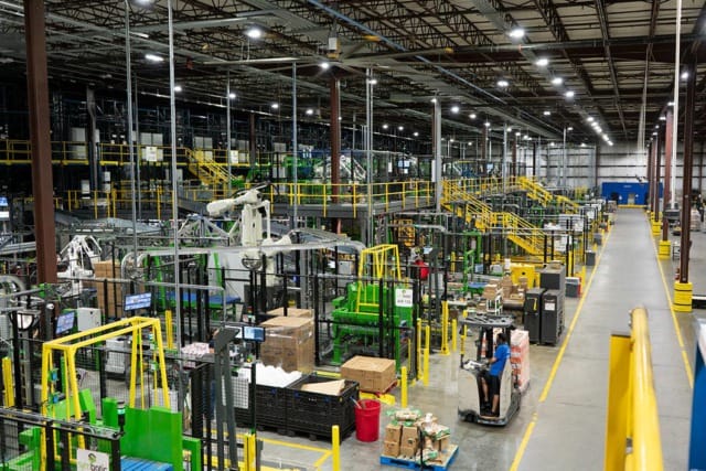 Vista panorámica del interior de una fábrica bien organizada, con maquinaria, cajas apiladas y un trabajador conduciendo una carretilla elevadora por el suelo.