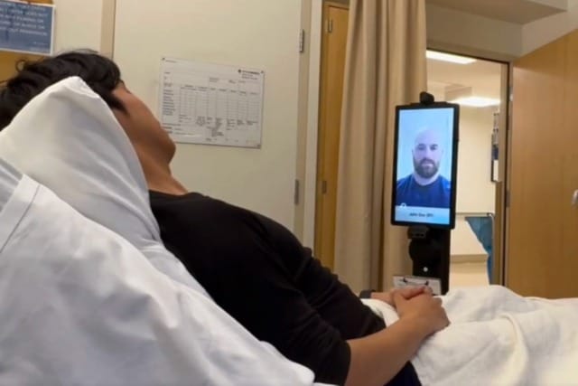 Un paciente en una cama de hospital mantiene una videollamada con un profesional sanitario a través de un dispositivo instalado en la pantalla de la habitación del hospital.