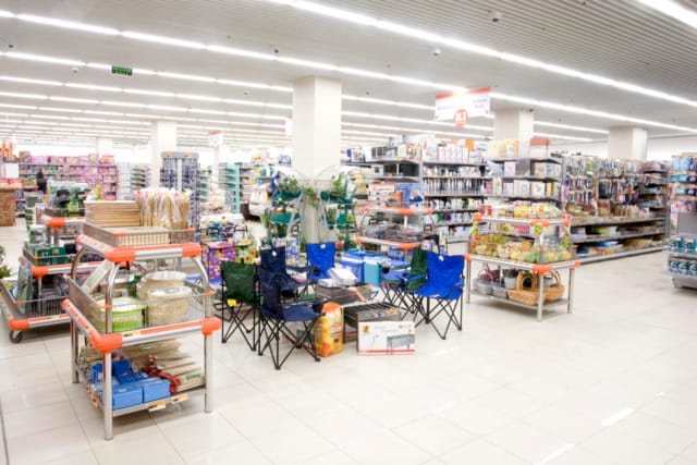 Un amplio pasillo de un supermercado con diversos artículos para el hogar, como sillas, cestas, productos de limpieza y productos para mascotas en las estanterías.