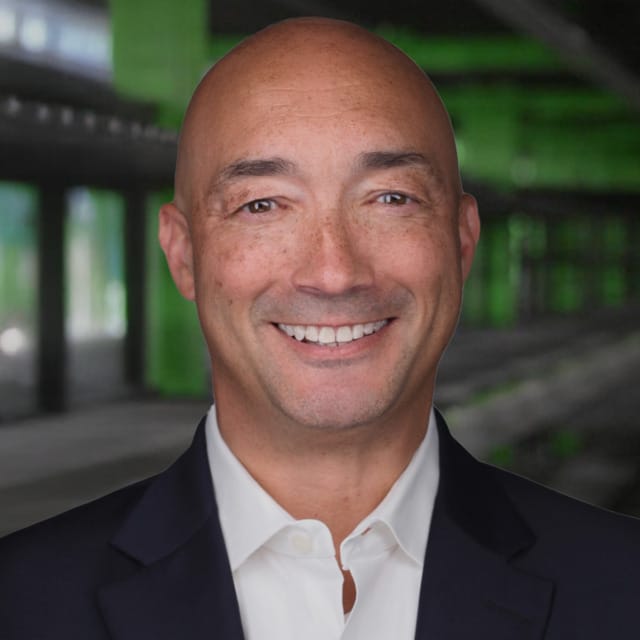 Hombre sonriente vestido con traje, de pie frente a un fondo arquitectónico moderno con elementos verdes y negros.
