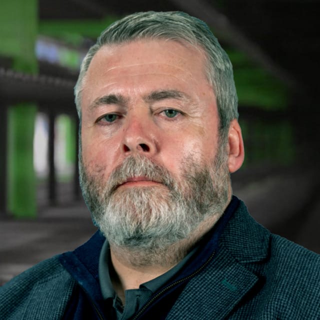 Hombre con barba y traje, de pie en un túnel verde con poca luz, con expresión seria
