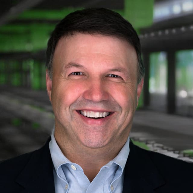 Hombre sonriente con traje oscuro sobre un fondo difuminado de color verde y negro.