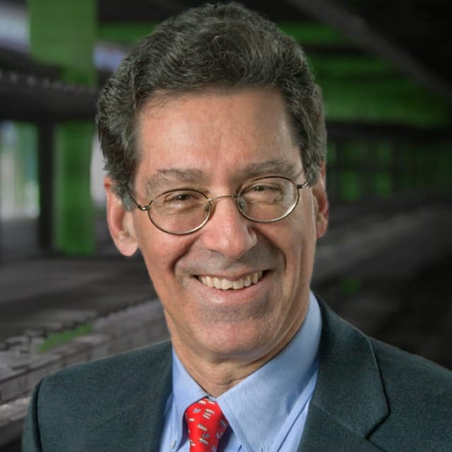Hombre sonriente con gafas, traje y corbata, de pie frente a un fondo borroso de color verde y gris.