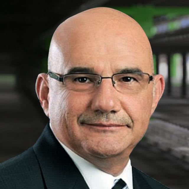 Un hombre calvo, con gafas y traje, de pie frente a un fondo borroso.