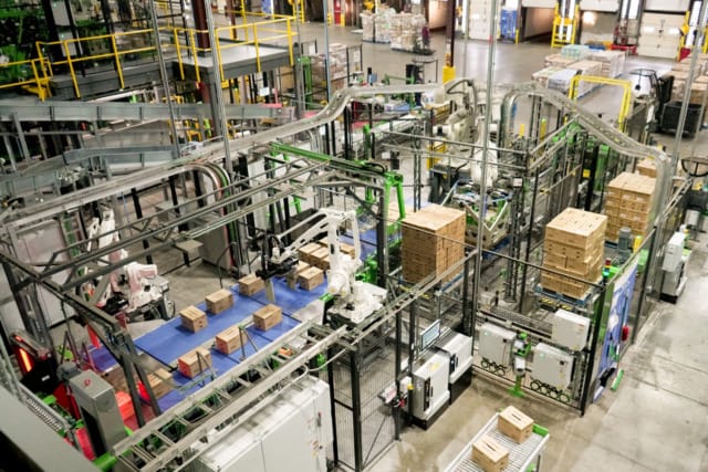Vista aérea de una instalación industrial automatizada con brazos robóticos que manipulan cajas en cintas transportadoras.