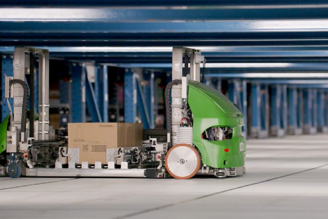 Carretilla elevadora automática de color verde transportando cajas en un gran almacén con estanterías metálicas azules al fondo.