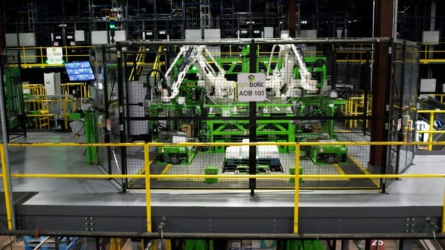 Brazos robóticos en un almacén de alta tecnología que gestionan el inventario dentro de una zona vallada con barandillas de seguridad amarillas.