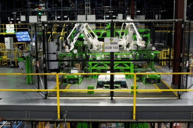 Brazos robóticos en un almacén de alta tecnología que gestionan el inventario dentro de una zona vallada con barandillas de seguridad amarillas.