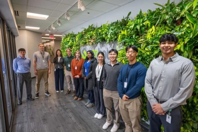 Un grupo de personas de pie en el pasillo de una oficina, con una pared vegetal verde, sonriendo y llevando tarjetas de identificación.