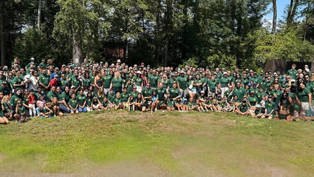 Foto de grupo de gran tamaño en la que aparecen personas con camisetas verdes a juego reunidas en una zona de césped con árboles al fondo.