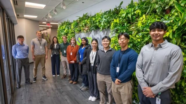 Un grupo de personas de pie en el pasillo de una oficina, junto a un muro vegetal vertical; todas llevan ropa de trabajo y tarjetas de identificación.