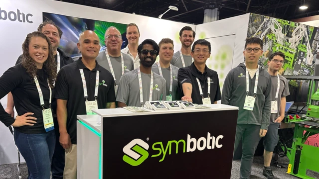 El equipo de Symbotic posa en el stand de una feria comercial, en el que se ven el logotipo de la empresa y los cordones identificativos; se trata de un grupo diverso de personas que sonríen.