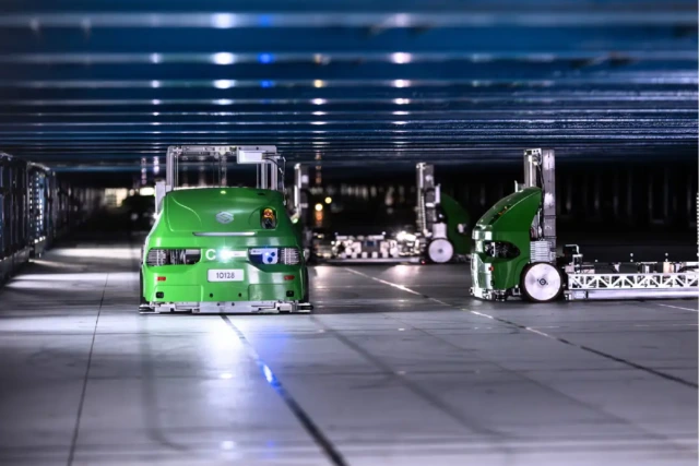 Automated green robots operate in a warehouse, moving quickly and efficiently under a structured roof.