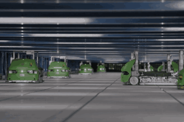 Những con robot màu xanh đang phân loại bưu kiện trong một kho hàng tự động mang phong cách tương lai, với những dãy kệ xếp chồng lên nhau ở phía sau.