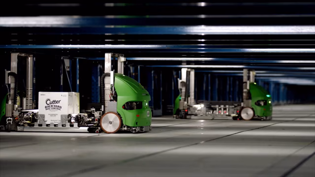 Những con robot tự hành màu xanh lá cây đang di chuyển trong một nhà kho, với những dãy kệ công nghiệp ở phía sau.