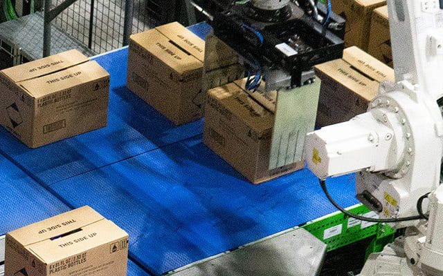 Cánh tay robot đang di chuyển các thùng carton trên băng chuyền trong kho.