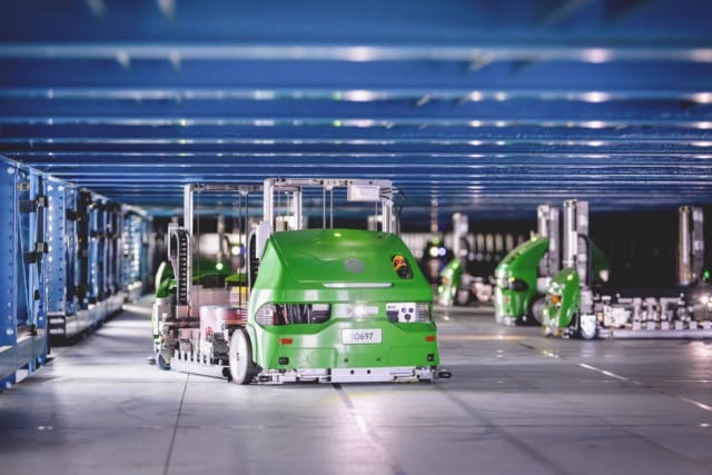 Những con robot kho tự động màu xanh lá cây di chuyển dưới khung kim loại màu xanh dương để quản lý hàng tồn kho tự động.