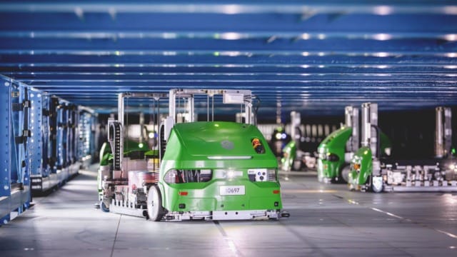 Những con robot tự hành màu xanh lá cây hoạt động trong môi trường kho hàng với các kệ hàng và dầm kết cấu màu xanh dương phía trên.