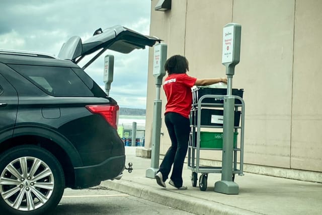 Một người mặc áo sơ mi đỏ đang chất đồ tạp hóa lên chiếc SUV màu đen tại điểm nhận hàng ngoài trời của cửa hàng.