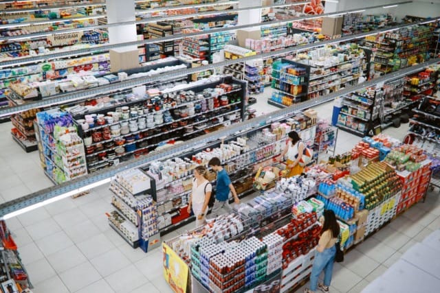 Mọi người đang mua sắm trong một lối đi rộng lớn của siêu thị, nơi các kệ hàng chất đầy đủ loại thực phẩm.