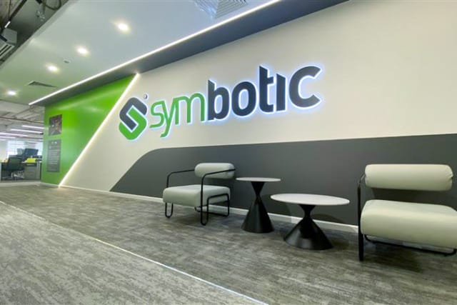 Khu vực chờ văn phòng hiện đại với ghế bành và bàn, tông màu xanh lá và trắng, cùng logo "Symbotic" trên tường.