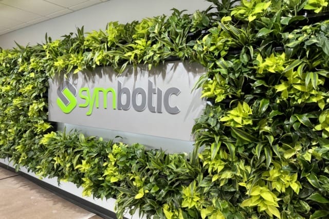 Mô hình vườn thẳng đứng với logo Symbotic ở giữa, được bao quanh bởi những cây xanh tươi tốt trong không gian văn phòng.