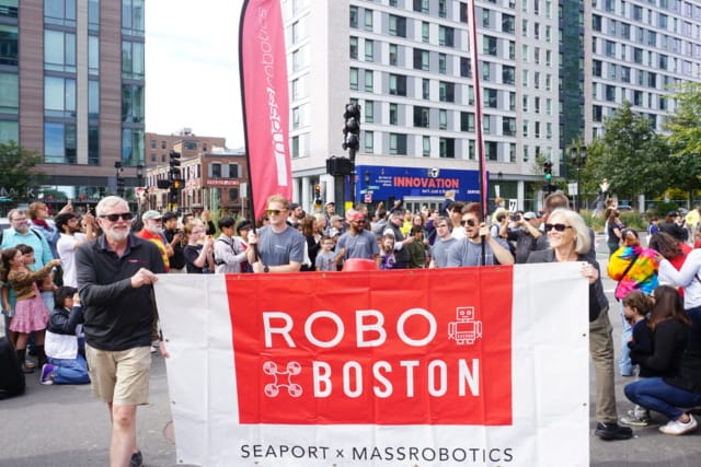 Crowd at Robo Boston event holding a large banner, with tall buildings and colorful signs in the background.